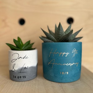 May include: Two small concrete planters with succulents. The planter on the left is white and grey with the text "Jade & Lee 24 09 15". The planter on the right is teal blue with the text "Happy 9th Anniversary 18/9".
