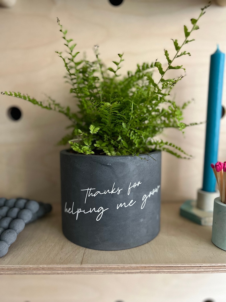 May include: A dark gray concrete planter with a green fern. The planter has the text "Thanks for helping me grow" in white script. A teal candle and a small container of matches are in the background.