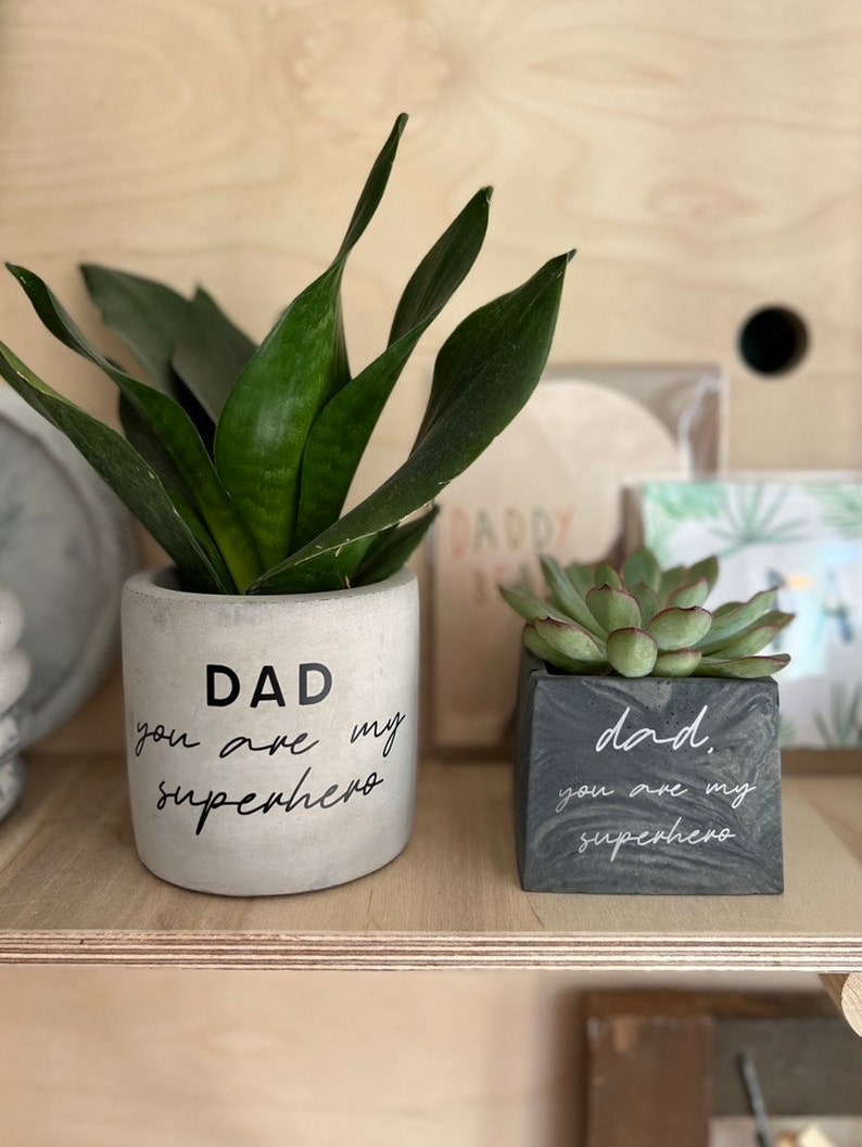 May include: Two concrete planters with plants. The larger, cylindrical planter has the text "DAD you are my superhero" in black. The smaller, square planter has the same text. The plants are green.
