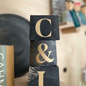 May include: Three stacked, dark gray concrete planters with gold lettering spelling "C & L". The top and bottom planters are square, while the middle one is cylindrical. The background includes a wooden shelf and other decorative items.