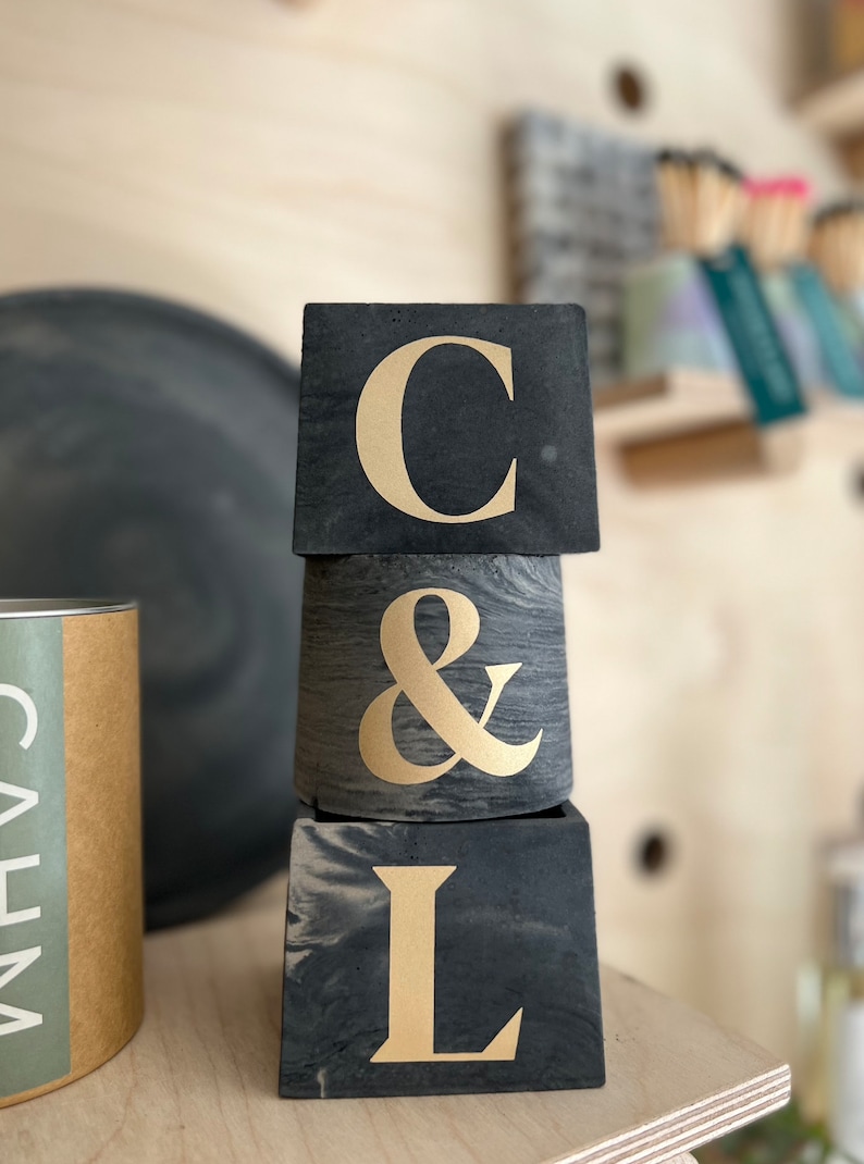 May include: Three stacked, dark gray concrete planters with gold lettering spelling "C & L". The top and bottom planters are square, while the middle one is cylindrical. The background includes a wooden shelf and other decorative items.