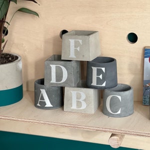 May include: A collection of small, gray concrete planters arranged on a wooden shelf. Each planter is a different shape and features a large, silver letter from A to F. The planters are various shades of gray.