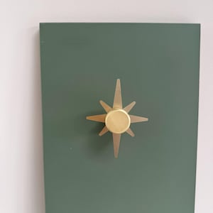May include: A gold-coloured starburst-shaped decorative knob is mounted on a dark green rectangular surface. The knob has a circular centre with eight radiating points. The background is a light-coloured wall.