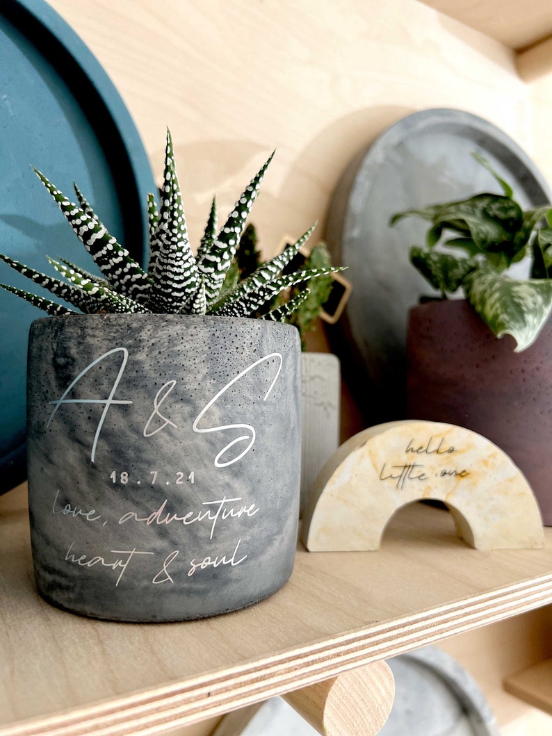 May include: A gray concrete planter with a succulent plant, featuring white script that reads "A & S", date, and "love, adventure, heart & soul". A small, arched decor piece reads "hello little one".