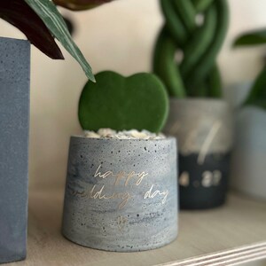 May include: A small, gray concrete planter with the words "happy wedding day" in gold script. The planter holds a green heart-shaped succulent. A small gold heart is below the text. The planter sits on a wooden shelf.