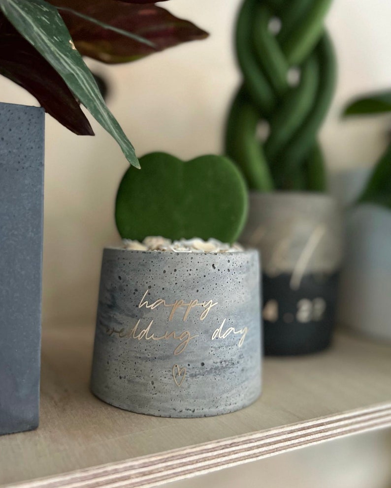 May include: A small, gray concrete planter with the words "happy wedding day" in gold script. The planter holds a green heart-shaped succulent. A small gold heart is below the text. The planter sits on a wooden shelf.
