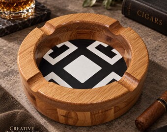 May include: A round wooden ashtray featuring a natural light brown wood grain finish and four integrated notches along the rim. The centre of the ashtray contains a circular black and white geometric pattern with a central square and four surrounding shapes.