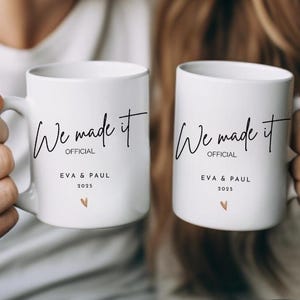 Peut inclure: Deux mugs en céramique blancs avec l'inscription "We made it OFFICIAL" en écriture noire. En dessous, les noms "EVA & PAUL", l'année "2025" et un petit cœur doré. Les tasses sont tenues par deux personnes.