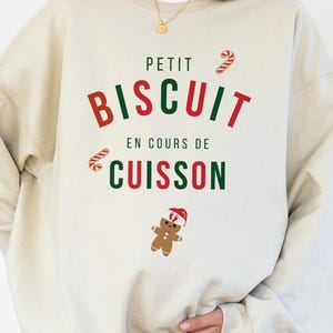Pode incluir: Camisola bege com o texto "PETIT BISCUIT EN COURS DE CUISSON" em vermelho e verde. O design inclui bengalas doces e um homem de gengibre com um chapéu de Pai Natal, perfeito para a época festiva.