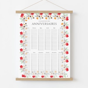 Op de afbeelding: Een bloemen verjaardagskalender print met de tekst "CALENDRIER DES ANNIVERSAIRES". De kalender is omlijst met rode, gele en witte bloemen en groene bladeren. De kalender hangt aan een houten balk.