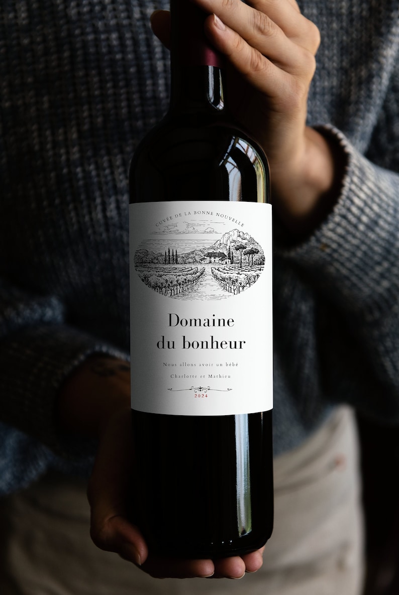 Puede incluir: Una botella de vino tinto oscuro con una etiqueta blanca con el texto "Domaine du bonheur" y una ilustraci&oacute;n de un vi&ntilde;edo. La botella est&aacute; sujeta por una persona que lleva un jersey gris. La etiqueta tambi&eacute;n incluye el texto "Nous allons avoir un b&eacute;b&eacute;" y el a&ntilde;o 2024.