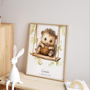 May include: A framed watercolor print featuring a cute hedgehog on a swing. The hedgehog is brown and tan, holding a small brown object. The print has a light wood frame and includes the name "Louise" and the date "21 septembre 2025".