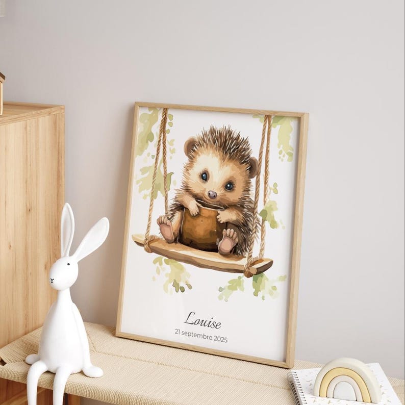 May include: A framed watercolor print featuring a cute hedgehog on a swing. The hedgehog is brown and tan, holding a small brown object. The print has a light wood frame and includes the name "Louise" and the date "21 septembre 2025".