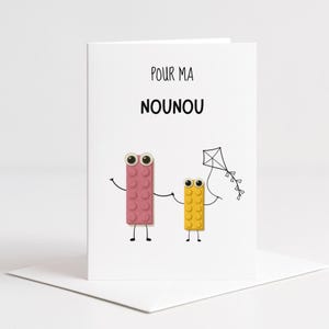 Può includere: Un biglietto di auguri bianco con la scritta "POUR MA NOUNOU" in alto. Il biglietto presenta personaggi a blocchi rosa e gialli che si tengono per mano, con un'illustrazione di un aquilone. Il personaggio a blocchi rosa è più alto di quello giallo.