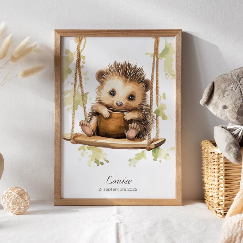 May include: A framed watercolor print featuring a cute hedgehog on a swing. The hedgehog is brown and tan with big blue eyes. The print includes the name "Louise" and the date "21 septembre 2025". The frame is made of light brown wood.