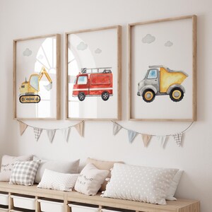 May include: Three framed watercolor prints featuring vehicles: a yellow excavator, a red fire truck, and a gray dump truck. Each print has a light wood frame and a white background with a few gray clouds. A garland of small flags hangs below the frames.