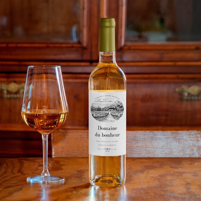 Puede incluir: Una botella de vino blanco y una copa de vino sobre una superficie de madera. La botella de vino tiene una parte superior verde y una etiqueta con las palabras "Domaine du bonheur" y una ilustraci&oacute;n de paisaje. La copa de vino est&aacute; llena de vino.