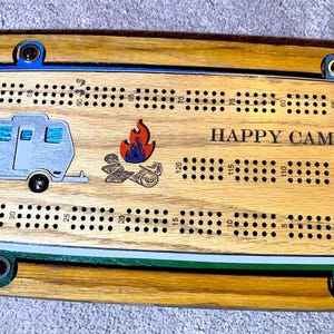 May include: A wooden cribbage board with a blue and white camper van illustration, a red and orange campfire illustration, and the text "HAPPY CAMPER".