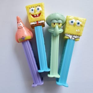 May include: A collection of four plastic Pez candy dispensers featuring characters from SpongeBob SquarePants. The set includes Patrick Star on a purple stem, SpongeBob on a blue stem, Squidward on a light green stem, and SpongeBob on a blue stem.