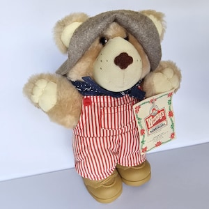 May include: A plush teddy bear wearing a gray hat, a blue bandana, and red and white striped overalls. The bear has tan fur and is holding a tag that says "Wendy's Hamburgers". The bear is wearing tan shoes.