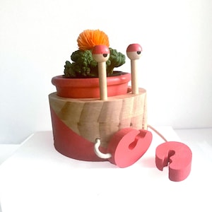 May include: A wooden crab-shaped planter with a small terracotta pot holding a green plant and an orange flower. The crab has red painted accents and two wooden antennae with red tips. The crab's claws are also red and connected by a white string.