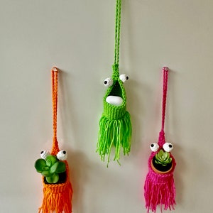 May include: Three colourful, handmade crochet plant holders with googly eyes hanging on a wall. The holders are orange, green, and pink, each with a different plant inside. The green holder has a white object inside.