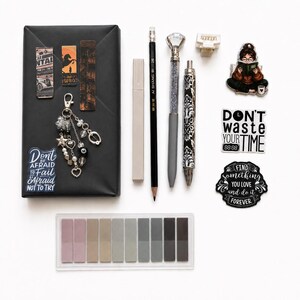 May include: A collection of stationery items including a black wrapped box with magnetic bookmarks, a beaded keychain, and a sticker reading Don't be afraid to fail, afraid not to try. Also includes a pencil, pens, a hair clip, stickers, and a set of adhesive tabs.