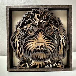 May include: A framed wood cutout of a dog's face. The dog has brown and white fur and is looking directly at the viewer. The frame is dark brown wood.