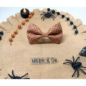 May include: A tan bow tie featuring a repeating pattern of small brown adhesive bandages. The bow tie is centered above a clear acrylic nameplate with the text "MERK & G" in black lettering, surrounded by black spider and orange bead accents.