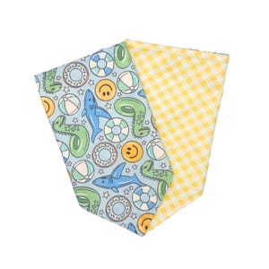 May include: A reversible pet bandana with a summer theme. One side features a light blue background with sharks, smiley faces, and pool floats. The other side has a yellow and white gingham pattern. The bandana is folded.