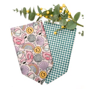 May include: Two dog bandanas. One is light pink with a pattern of flamingos, rainbows, smiley faces, beach balls, and seashells. The other is teal with a white checkered pattern. Greenery with yellow berries is in the background.
