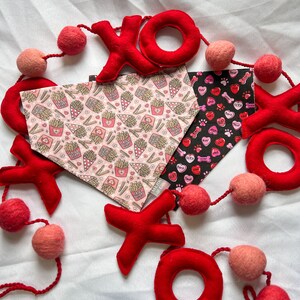 May include: Red felt XOXO garland with pink pom poms and two dog bandanas. One bandana has a pink and white pattern with hearts and french fries. The other bandana has a black background with pink hearts, paw prints, and bones.