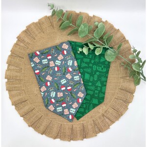 May include: Two dog bandanas with fishing and camping themes. One is gray with fishing tackle and bobbers, the other is green with camping equipment. They are displayed on a burlap placemat with a ruffled edge, with a sprig of greenery.