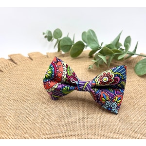 May include: A colorful bow tie with a paisley pattern in shades of pink, orange, purple, green, and blue. The bow tie is made of fabric and has a classic bow shape.