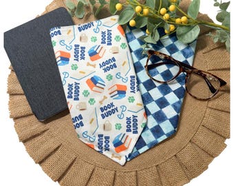 May include: Two triangular fabric bandanas, one white with a repeating pattern of books, coffee mugs, glasses, and the text "BOOK BUDDY," and one with a blue and white checkered pattern, accompanied by a pair of brown-rimmed eyeglasses.