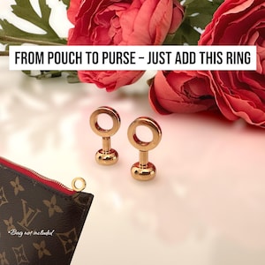 May include: Two gold rings with a circular design and a small base, next to a brown pouch with a red zip. The text "FROM POUCH TO PURSE - JUST ADD THIS RING" is displayed above the rings. Red flowers are in the background.