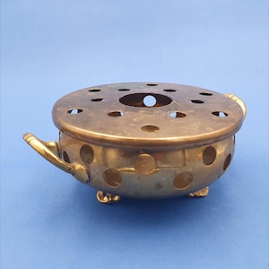 Op de afbeelding: Een vintage messing wierookbrander met een geperforeerd deksel en een ronde behuizing. De brander heeft kleine voetjes en handvatten aan weerszijden. Het messing heeft een aangetaste, verouderde uitstraling, wat suggereert dat het antiek is.