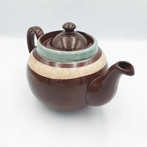 Può includere: Una teiera marrone con un design a strisce marroni chiare e blu. La teiera ha un coperchio e un beccuccio.