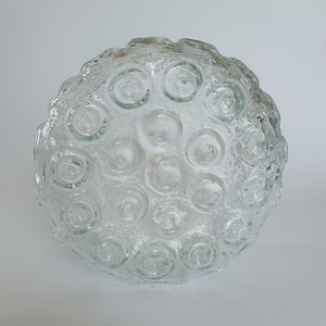 May include: A clear glass decorative bowl with a textured surface and a pattern of circular indentations. The bowl is round and appears to be made of thick glass. The bowl is sitting on a white surface.