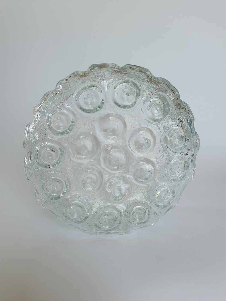 May include: A clear glass decorative bowl with a textured surface and a pattern of circular indentations. The bowl is round and appears to be made of thick glass. The bowl is sitting on a white surface.