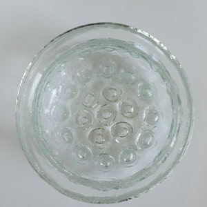 May include: Clear glass flower frog with a circular design. The frog has a textured rim and a central area with multiple circular openings. The glass has a slightly frosted appearance, and the overall shape is round.
