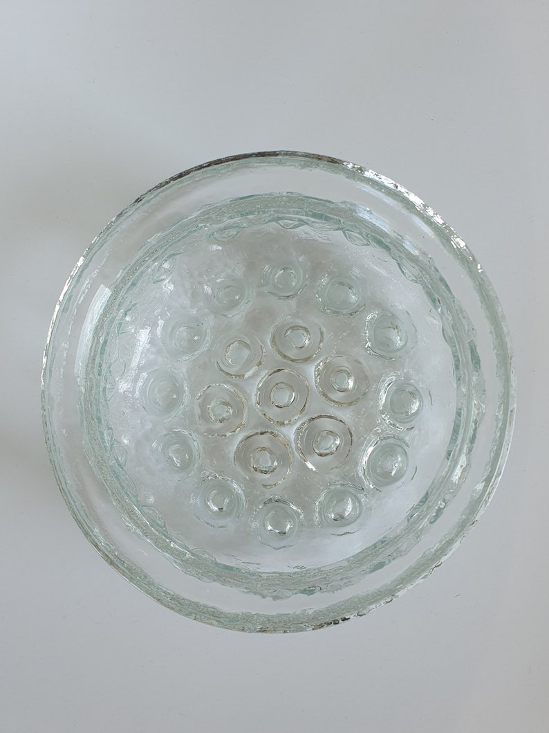 May include: Clear glass flower frog with a circular design. The frog has a textured rim and a central area with multiple circular openings. The glass has a slightly frosted appearance, and the overall shape is round.