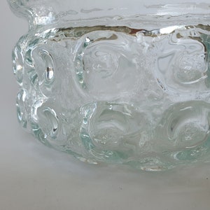May include: A clear glass bowl with a textured surface and a series of circular indentations around the sides. The bowl has a wide, open top and a slightly flared rim. The glass appears to be thick and heavy, with a slightly frosted appearance.