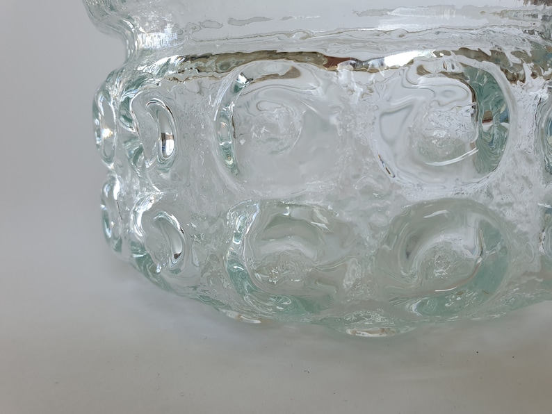 May include: A clear glass bowl with a textured surface and a series of circular indentations around the sides. The bowl has a wide, open top and a slightly flared rim. The glass appears to be thick and heavy, with a slightly frosted appearance.