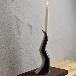 May include: A contemporary candle holder with a lit, cream-coloured taper candle. The holder is black and silver, placed on a brown book. The book is on a light wood surface, against a textured, off-white wall.
