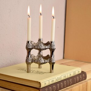 May include: A silver-toned, three-candle holder with lit white candles. The decorative candle holder sits atop a stack of books with the text "ERDKREIS 1952" visible. The background is a light pink wall and a wooden surface.