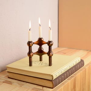 May include: A bronze candle holder with three lit white candles sits atop two vintage books. The candle holder has a geometric design and is placed on a light-colored book with a darker brown book underneath.