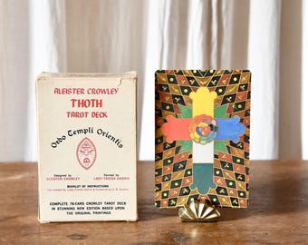 Puede incluir: Un juego de cartas de tarot vintage con una caja con el texto "Aleister Crowley Thoth Tarot Deck" y una carta colorida con un diseño de cruz. El juego incluye un folleto de instrucciones y se muestra sobre una superficie de madera.