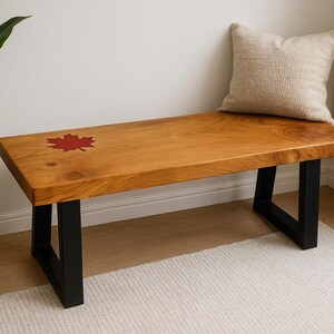 May include: A wooden bench with a red maple leaf design on the top. The bench has a natural wood finish and black metal legs. A beige pillow rests on the bench. A potted plant is to the left.