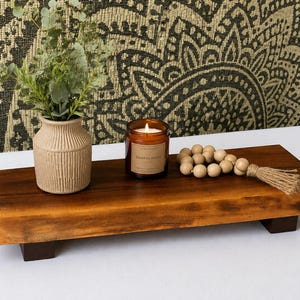 Puede incluir: Una bandeja de madera marrón oscuro con un pequeño jarrón beige que contiene vegetación, una vela encendida en un frasco de vidrio y una hebra de cuentas de madera con una borla de yute. La bandeja es rectangular y se apoya en cuatro patas pequeñas de color marrón oscuro.